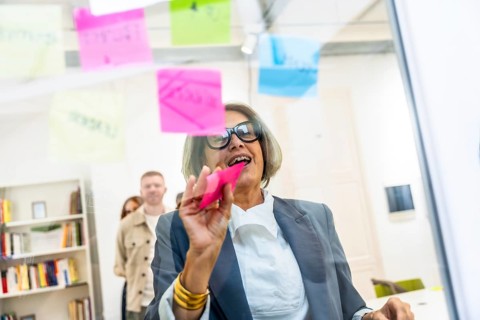 Mature businesswoman attaching colorful sticky notes on a clear glass board, collaborating on