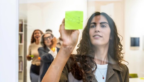 Young businesswoman sticking a green sticky note on a transparent glass wall during a team meeting,