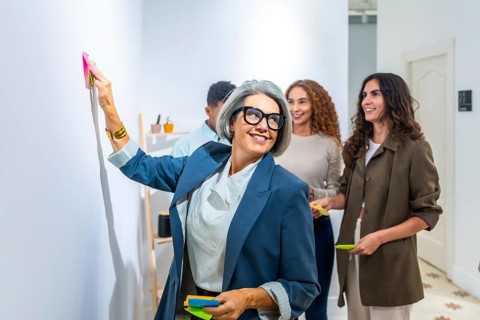 Confident gray haired businesswoman in glasses posts a sticky note during a lively team