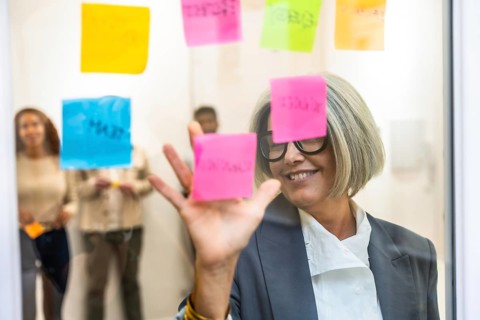 Senior business woman smiling, organizing colorful sticky notes on a glass whiteboard during a
