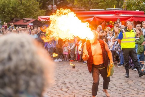 A fire-breather entertains the crowd during an outdoor performance, flames shoot out of his mouth,