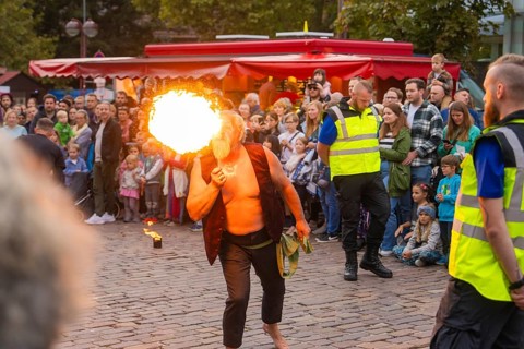 A fire eater entertains a crowd on a cobbled street in front of red stalls, Fire Night,