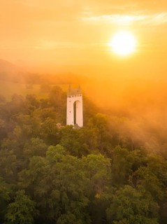 White tower rises out of misty forest while the sun illuminates orange sky, Schönbergturm,