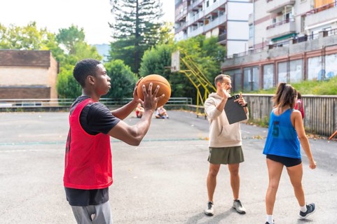 Young basketball player practicing shooting technique on outdoor court, receiving guidance from