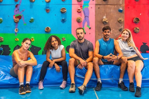 Five athletes resting on a vibrant blue crash pad in a colorful bouldering gym, enjoying a well