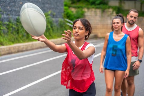 Young woman wearing a red training bib practices passing a rugby ball on a sports field, while