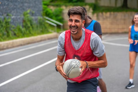 Male rugby player wearing a red training bib smiles broadly while holding the ball during practice