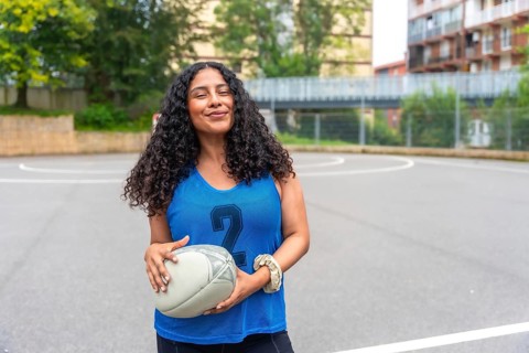 Female rugby player wearing a blue shirt, smiling confidently while holding a rugby ball on an