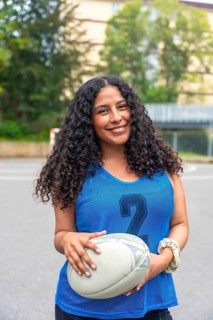 Confident female rugby player is smiling while holding a rugby ball, standing on a sports field,