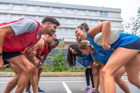Two rugby teams, one in red and one in blue, are facing each other and smiling before a match, with
