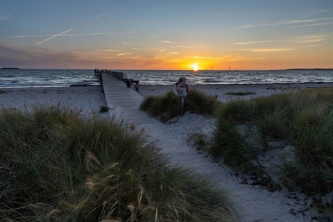 Sunset sky over the sea, wooden walkway and lifebuoy, framed by beach grass and calm water, Kobaek