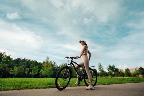 A woman stands beside her bicycle in a lush green park. She wears comfortable athletic clothing and