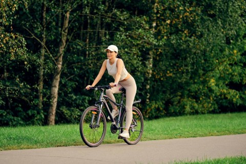 A slender woman is riding her bicycle on a smooth pathway bordered by lush greenery. She is dressed
