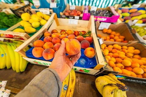 A hand holding a peach above a market stall with various fruits such as bananas and peaches,