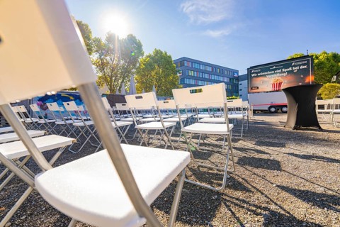 White chairs lined up outside in sunny weather in front of a building and a large screen, open air