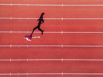 Aerial view of a female individual (runner) on a red athletics track (tartan track) with shadow as