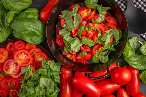 A pan filled with sliced tomatoes and basil leaves, surrounded by fresh vegetables
