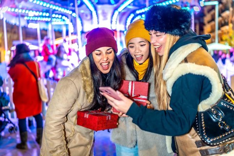 Three cheerful young women holding christmas gifts and using a smartphone in a christmas market