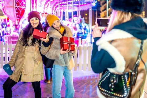 Three happy female friends exchanging christmas gifts and taking photos in front of a carousel at a