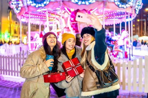 Three cheerful young women taking a selfie at a christmas market, holding gifts and warm drinks,