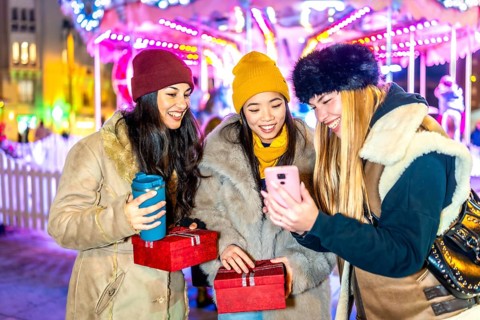 Three happy women using a smartphone and holding christmas gifts at night in a christmas market