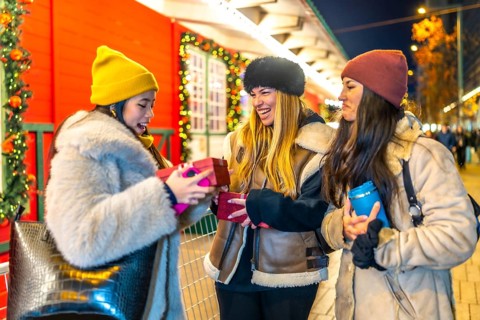 Three cheerful young women exchanging christmas presents at a decorated outdoor night market,