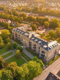 Large building with flat roof and balconies, surrounded by trees and streets in a sunny