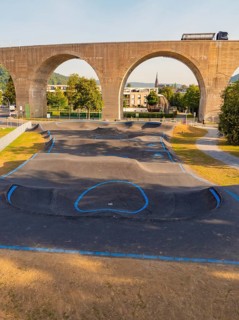 A pump track under a bridge with concrete arches and green surroundings, pump track, Nagold, Black