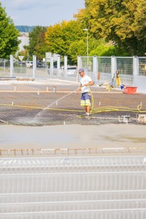 Construction worker watering concrete on a building site, surrounded by safety fences, car park
