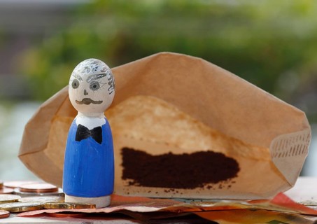Male wooden cone figure with coins and banknote in front of filter bag with coffee North