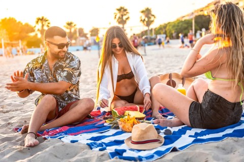 Three friends relaxing on a beach towel, sharing fresh fruit like watermelon and pineapple while