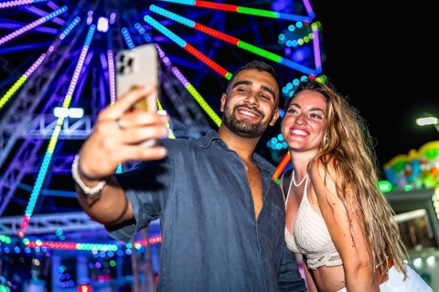 Young couple smiling and taking a selfie with smartphone in front of a colorful ferris wheel at an