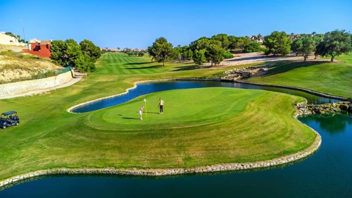 Two golfers are putting on a green surrounded by water, with lush fairways and trees in the