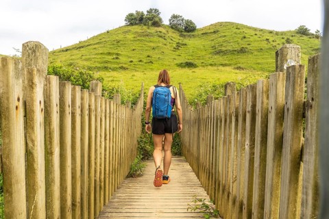 Female hiker with backpack walking on wooden footbridge leading towards a green hill in zumaia,