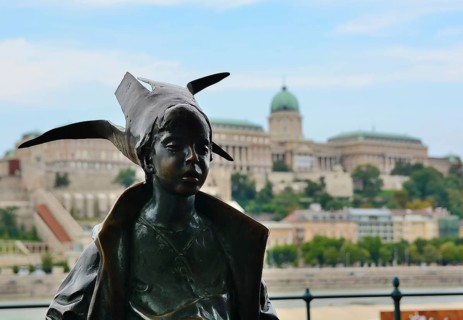 Bronze statue of a little princess wearing a bird shaped hat, overlooking the majestic buda castle