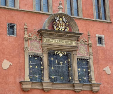 Architectural detail showcasing an ornate window with a coat of arms, latin inscription, and