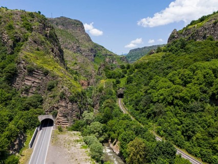 Road tunnel and railway tunnel through forested mountain landscape under blue sky, aerial view,