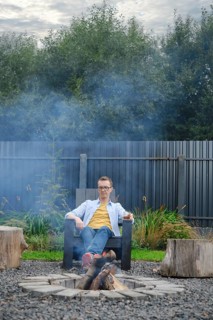 A middle-aged man sits comfortably in a chair next to a campfire surrounded by logs. Smoke rises