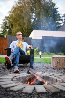 A retired man sits comfortably in a wooden chair by a crackling fire pit in his backyard. He enjoys