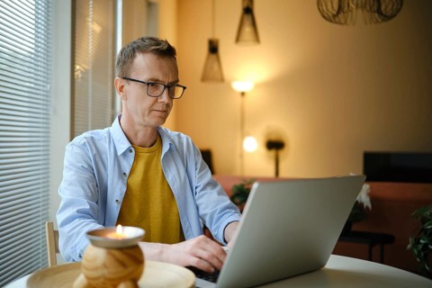 A middle-aged man sits at a table and typing letter on his laptop in a living room. Soft lighting