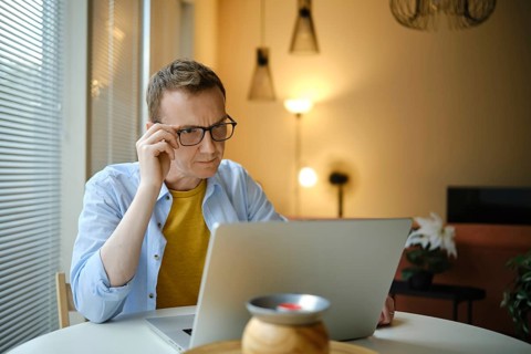 A focused man with glasses is working on his laptop with financial documents at home in the evening
