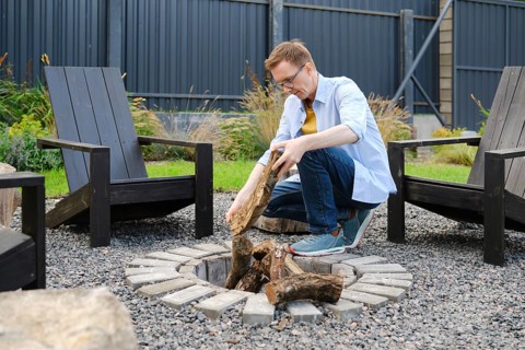A middle-aged man is kneeling beside a circular stone fire pit, carefully placing logs into the