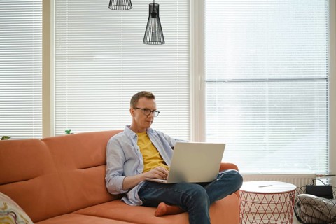 A man is seated on an orange couch, focused on his work on a laptop. Sunlight streams through the