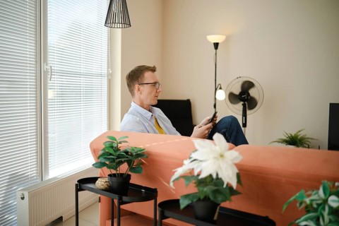 A man sits comfortably on a couch in a modern living room with large windows and natural light. He