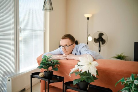 A middle-aged man leans on a colourful couch, gazing thoughtfully at potted indoor plants nearby.