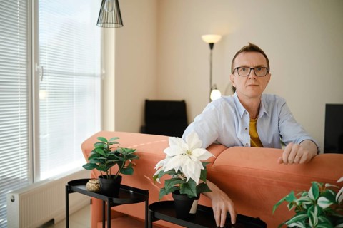 A middle-aged man sits on an orange couch in a contemporary living room. He appears thoughtful as