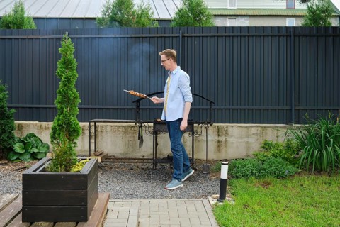 In a vibrant backyard garden, a man stands next to a grill, skillfully cooking skewered food. Lush