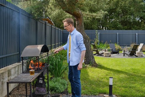 A man stands beside a barbecue grill, actively tending to the flames in a vibrant backyard. The