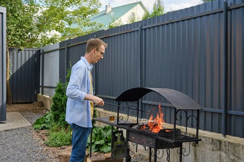 A man stands by a grill in a backyard, tending to the flames. The surrounding area features