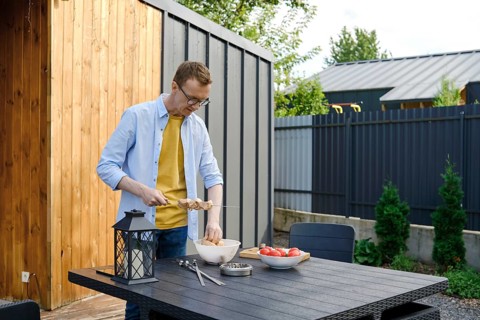 A man wearing a yellow shirt and denim jacket is preparing meat skewers at a wooden table in a